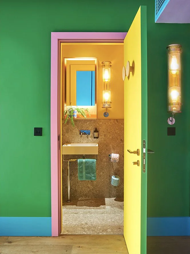 Colorful bathroom door leading to a vibrant room with green, pink, yellow, and blue walls, illuminated wall sconces, and a small sink with a green towel.