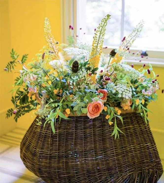 A wicker basket filled with colorful flowers and greenery, placed on a yellow surface near a window.