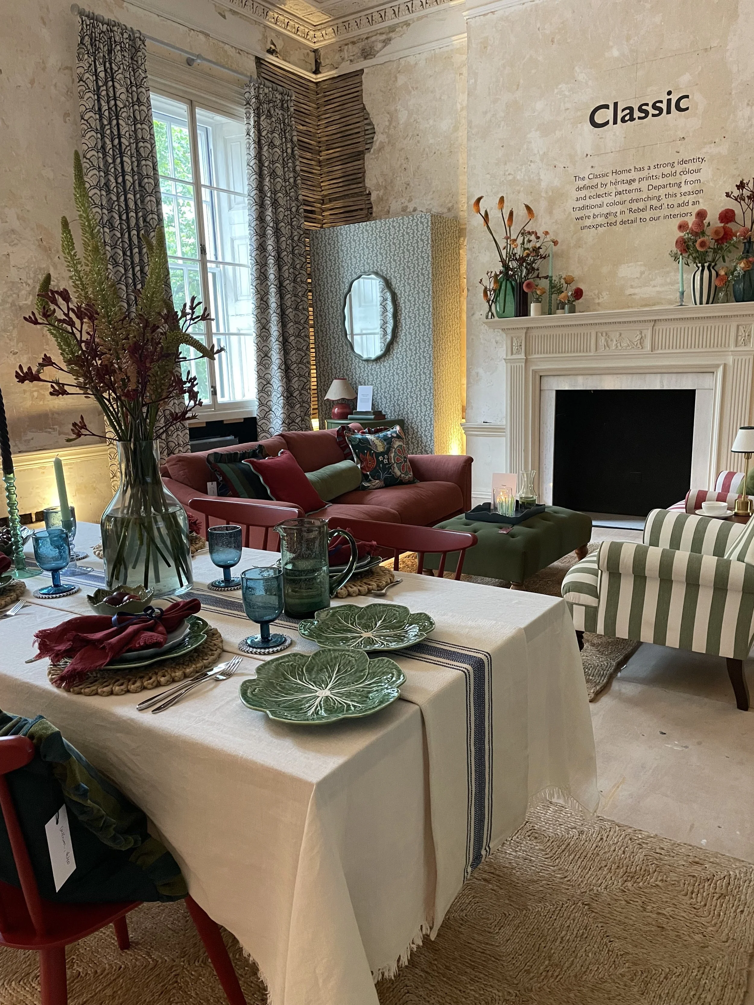 A living room decorated with vintage and classic furniture, including a fireplace, a striped armchair, a red sofa, and a dining table set with green plates, blue glasses, and floral arrangements.
