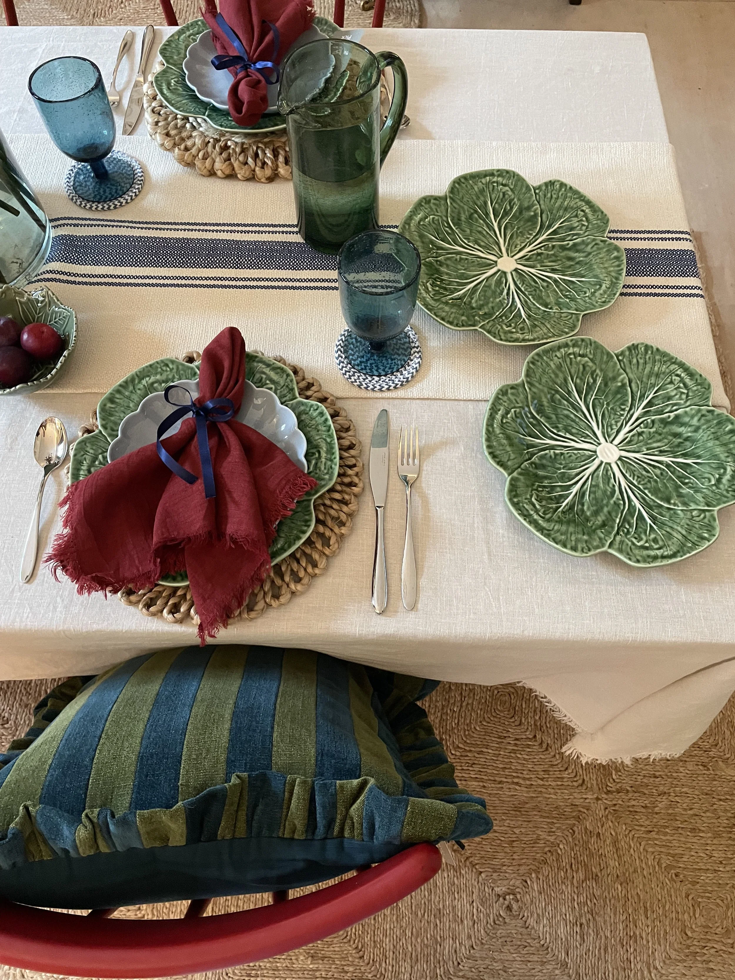 A dining table set with green leaf-shaped plates, red cloth napkins tied with dark ribbon, blue glasses, a green pitcher, and a bowl of grapes, on a white tablecloth with a striped navy and white runner.