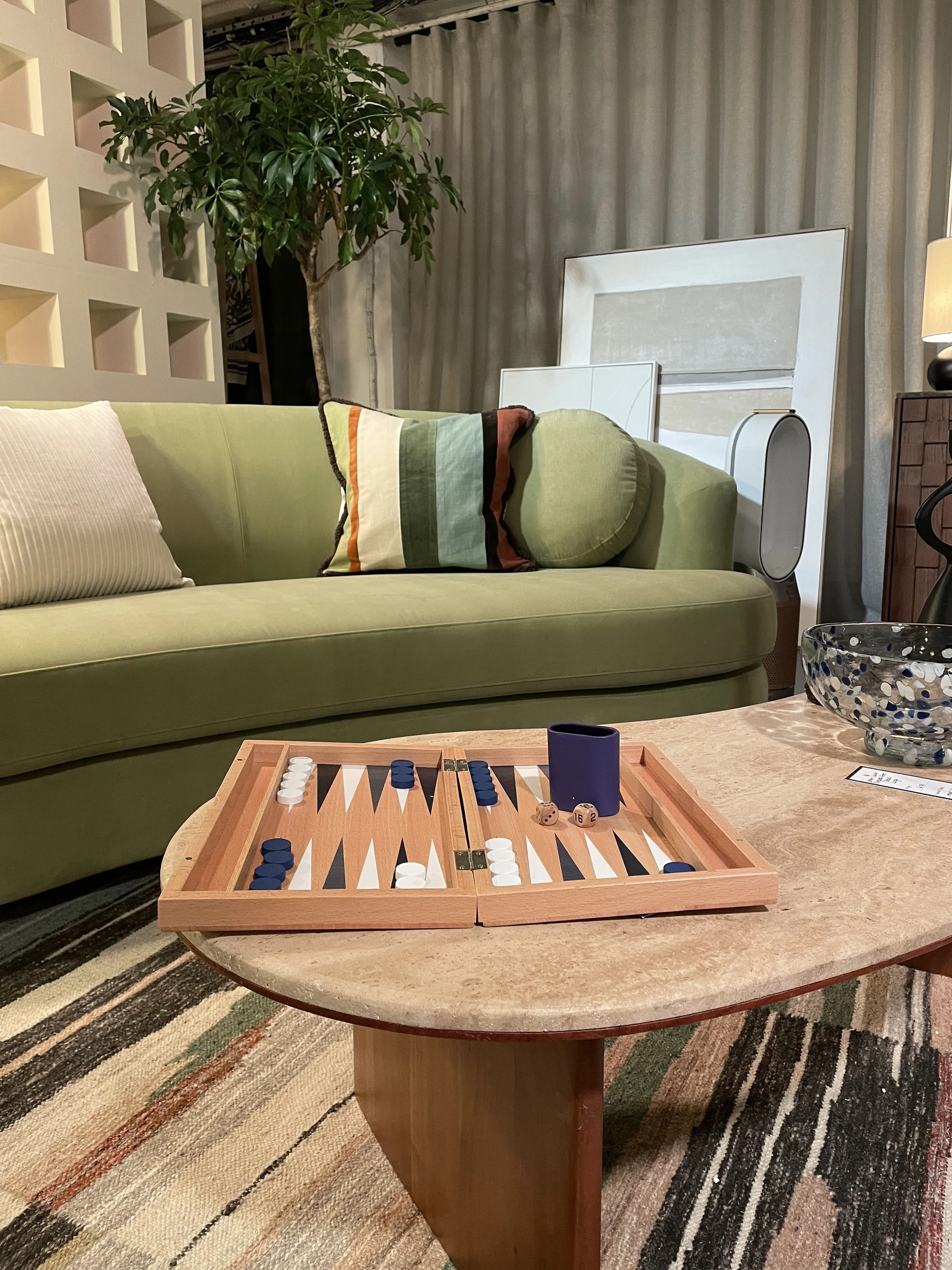 A living room with a green sofa, a striped pillow and a round bolster, a wooden coffee table with a backgammon game, and artwork against a curtain backdrop.