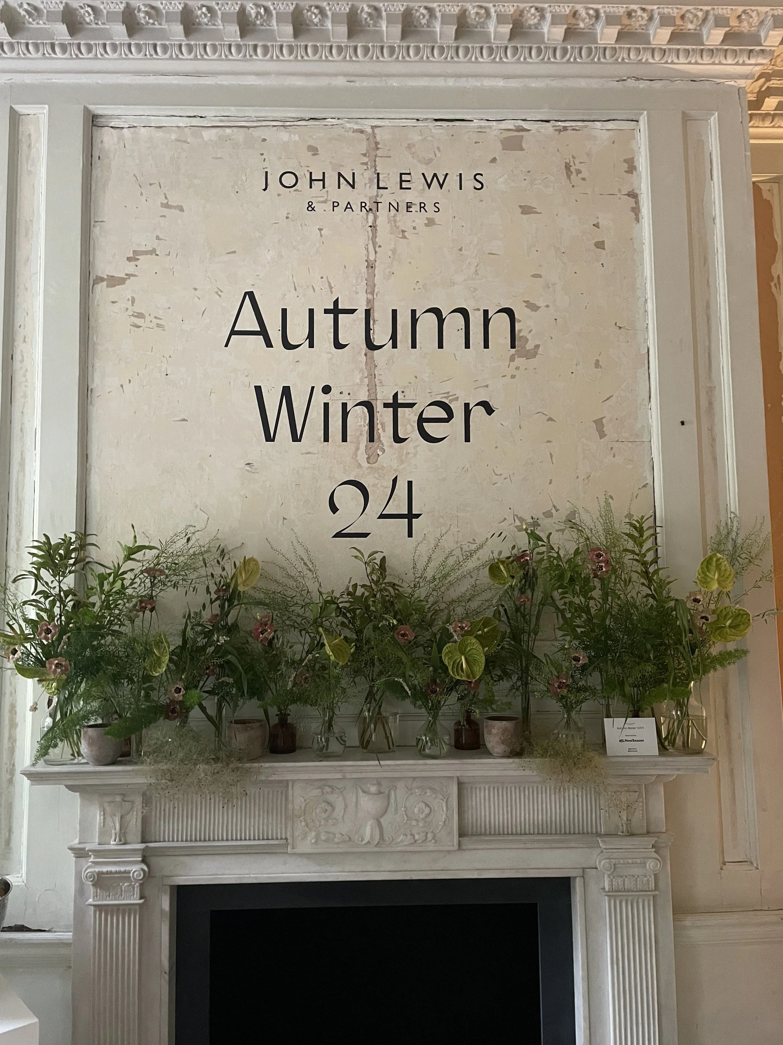 A large sign above a decorated fireplace reads "John Lewis & Partners Autumn Winter 24". The mantel is decorated with green foliage and flowers in various small vases.