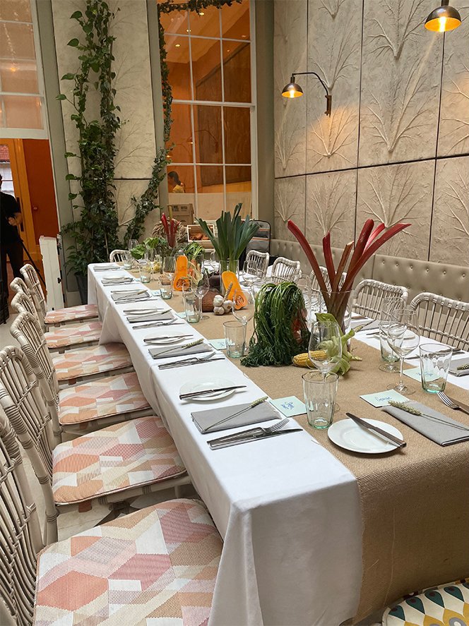 A long dining table set for a meal in a well-lit room with large windows, decorated with greenery and tropical plants as centerpieces, with place settings including plates, glasses, and utensils, and along the sides are patterned chairs.