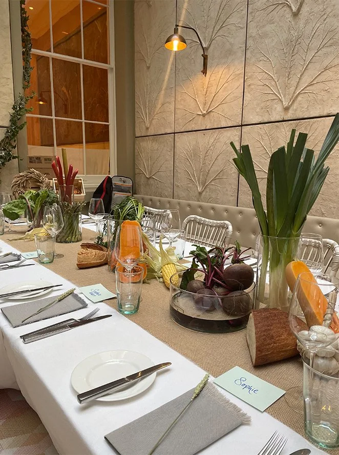 A decorated dining table with a beige burlap runner, glass vases with large green leaves, bowls of root vegetables, slices of bread, and place cards, set for a meal in a cozy room with textured wall art and a warm wall-mounted light fixture.