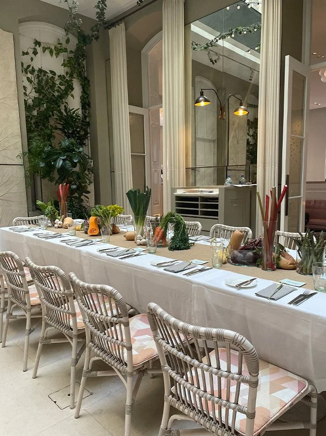 A long dining table set for a meal with plates, silverware, and glasses, decorated with vegetables and greenery, in a bright, elegant room with large windows, greenery, and soft lighting.