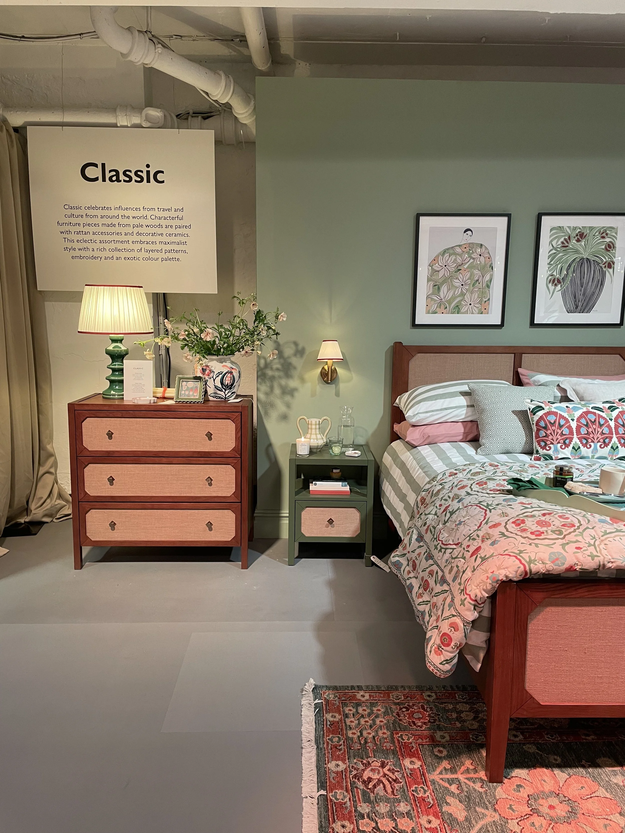 A bedroom display with a green accent wall, framed artwork featuring floral and geometric patterns, a wooden bed with a floral quilt, pink and patterned pillows, a bedside table, a chest of drawers with a lamp, a vase with flowers, and a small wall s