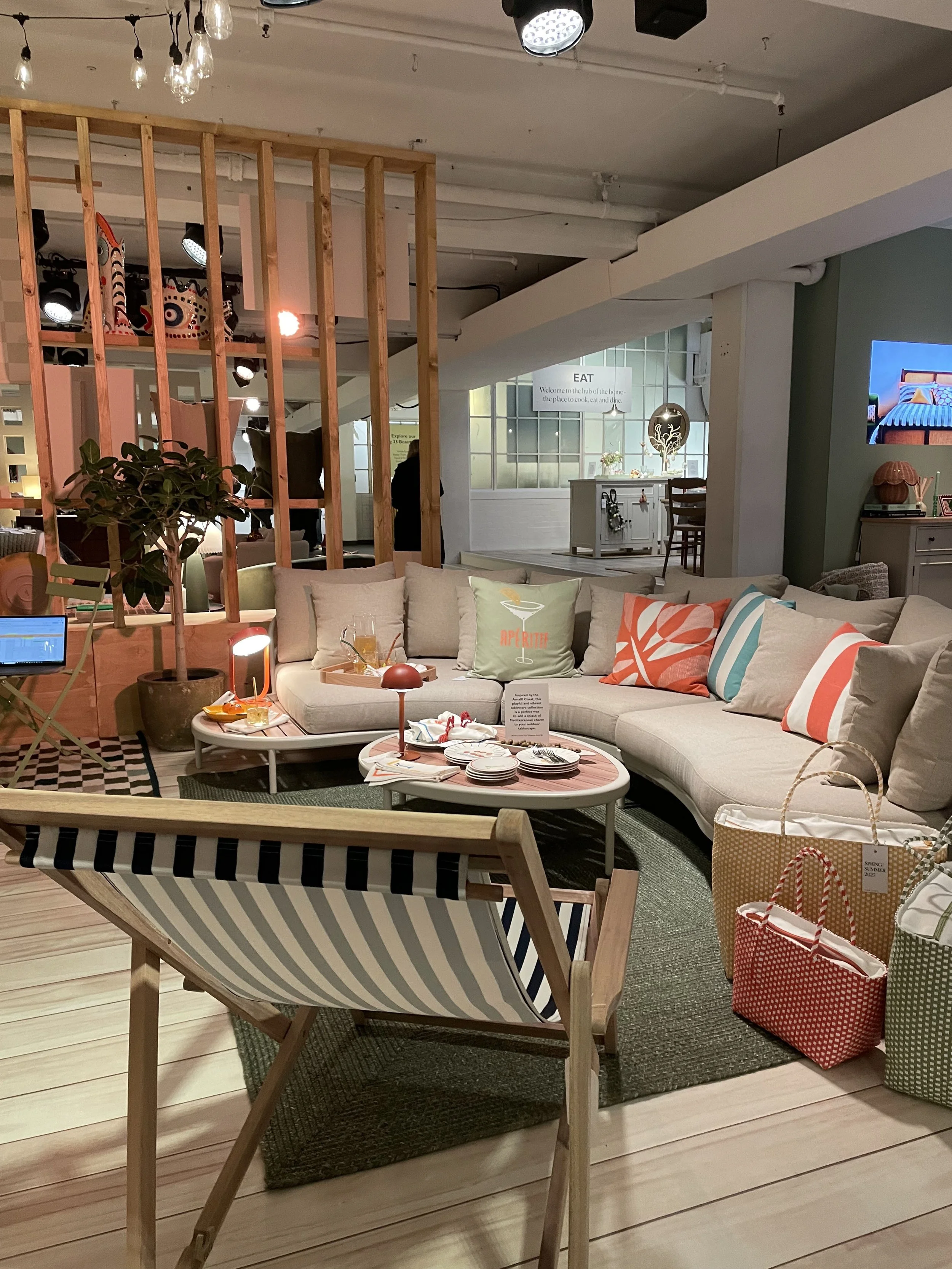 Stylish living room with a large beige sectional sofa, colorful pillows, a round coffee table with dishes, and a striped lounge chair. Decor includes a potted plant, various bags, and background lighting and art, creating a cozy, modern ambiance.