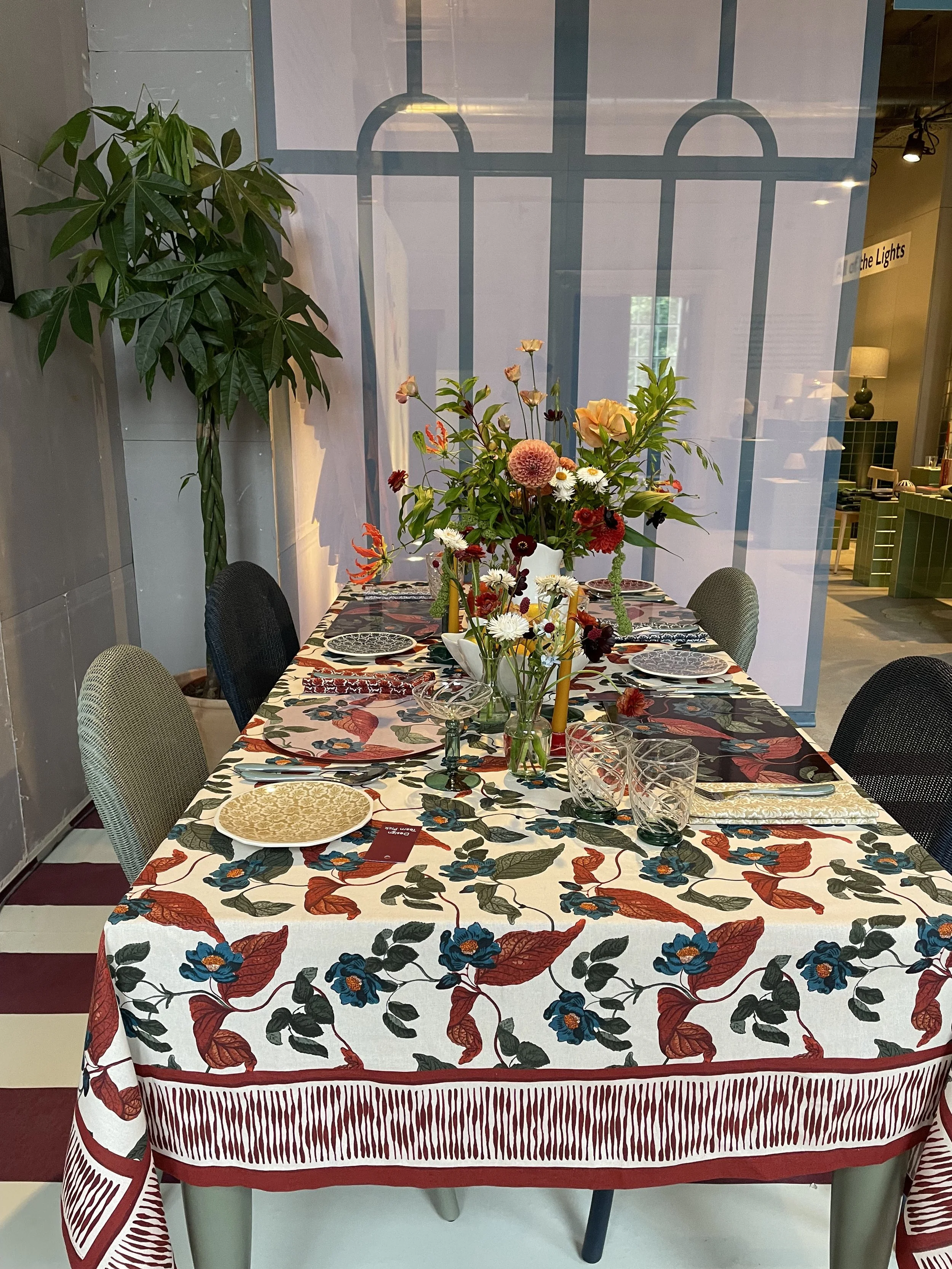 A dining table decorated with a colorful floral tablecloth, set with plates, glasses, and silverware, and topped with a large floral centerpiece, situated in a room with a large plant and a glass partition.