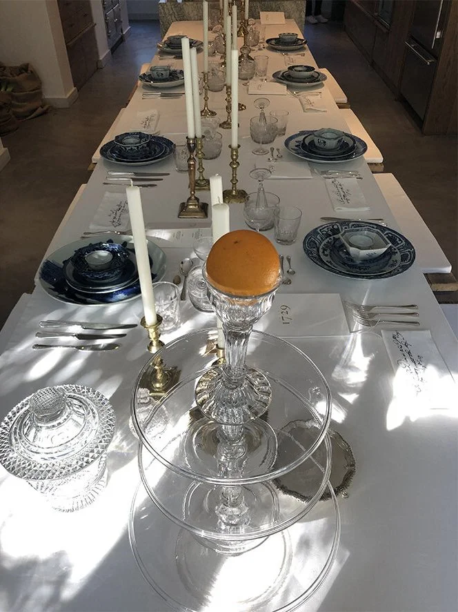 A long dining table set with blue and white china, glassware, silverware, white candles in brass candle holders, and a cake on a glass pedestal centered on the table. The table is in a well-lit room with sunlight casting shadows.