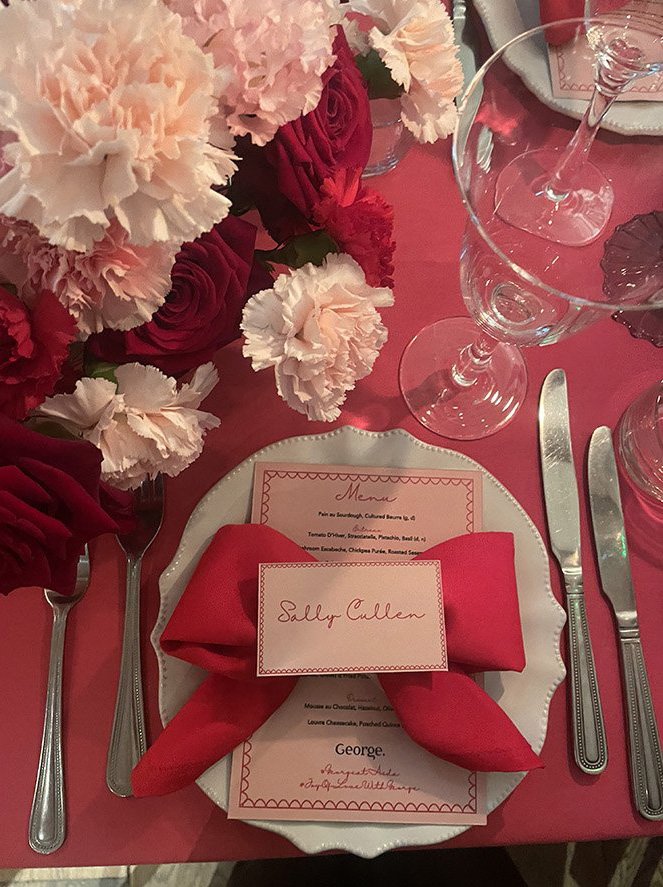 A place setting at a formal event featuring a white decorative plate, a red cloth napkin folded with a personalized name card reading 'Sally Cullen,' silverware, a clear glass goblet, and a floral centerpiece with pink, red, and white flowers on a pi