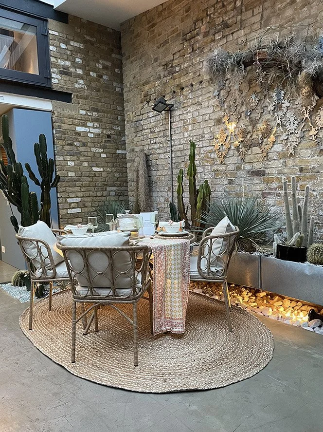 Indoor dining area with a round table, six chairs, a patterned tablecloth, and decorative plants including cacti and succulents on a stone and brick wall. The area features a circular rug and a small landscaped section with rocks and trailing plants.