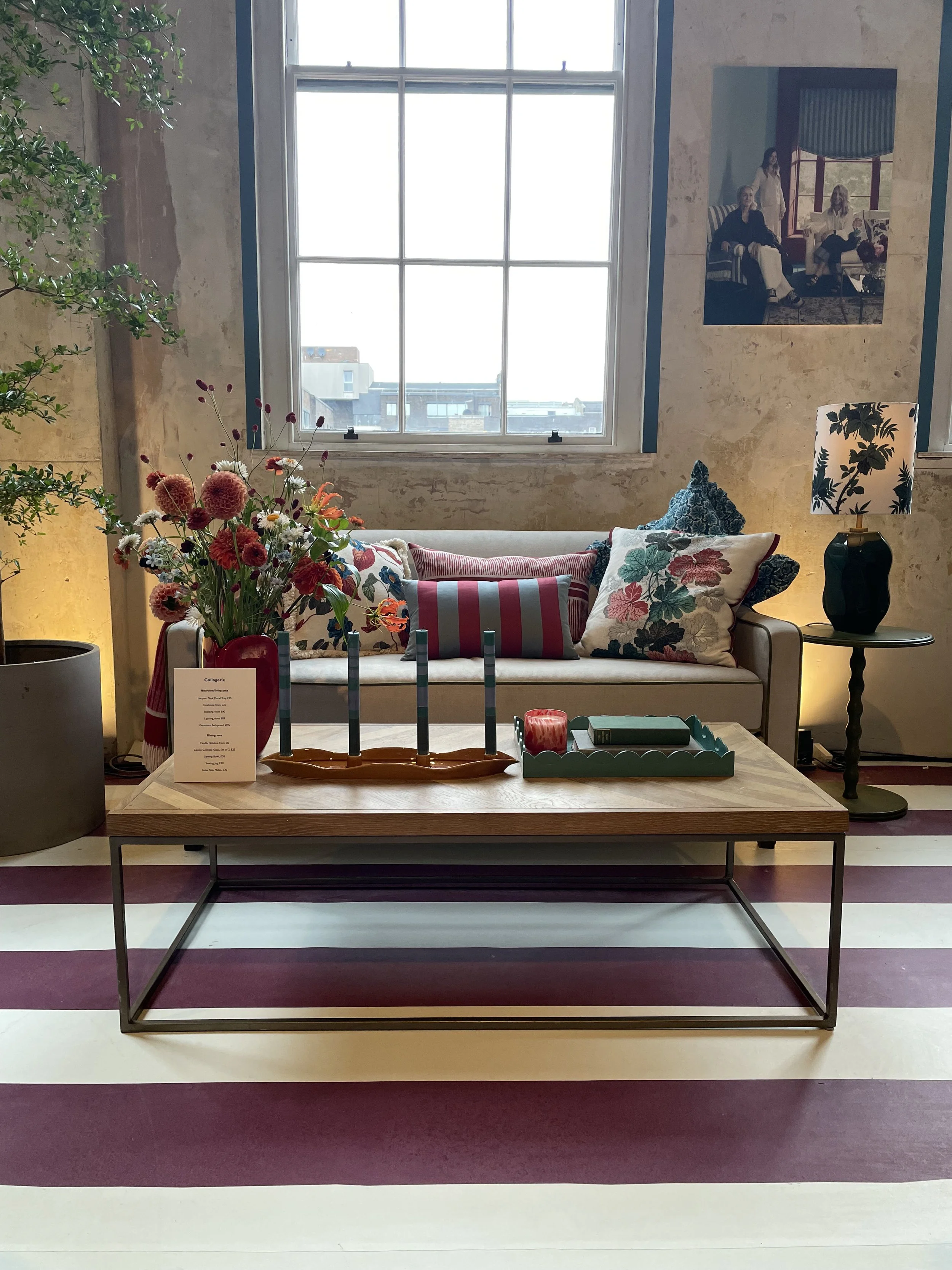 A cozy living room corner with a beige sofa adorned with patterned cushions, a wooden coffee table with a flower arrangement, candles, and books, a window in the background, a black side table with a lamp, a large potted plant, and a framed photograp