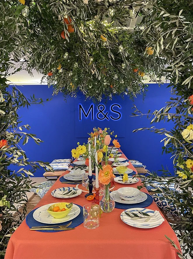 A decorated dining table set with plates, bowls, cutlery, and colorful flowers, surrounded by greenery and orange fruit decorations, with a blue wall and 'M&S' sign in the background.