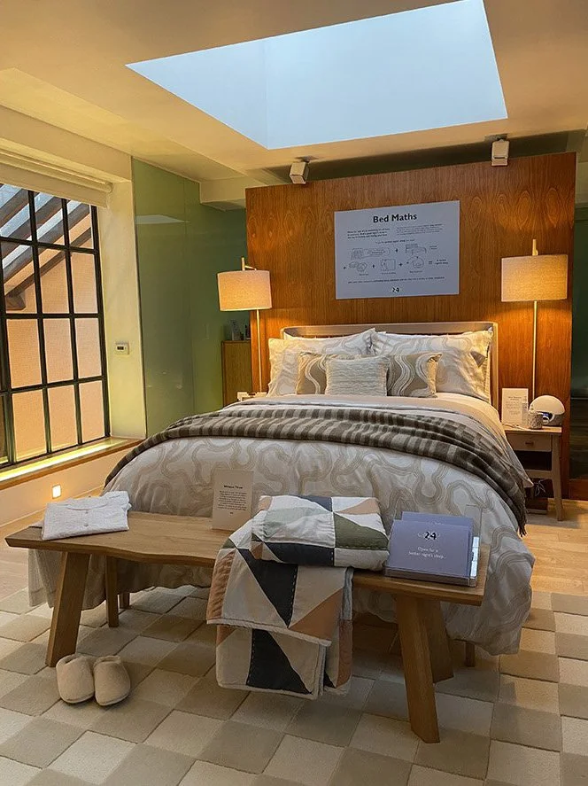 A neatly made bed with decorative pillows and a beige comforter in a hotel room. On the wall above the bed is a sign titled 'Bed Maths.' There are two bedside lamps, one on each side, and a wooden bench at the foot of the bed with towels, a folded bl