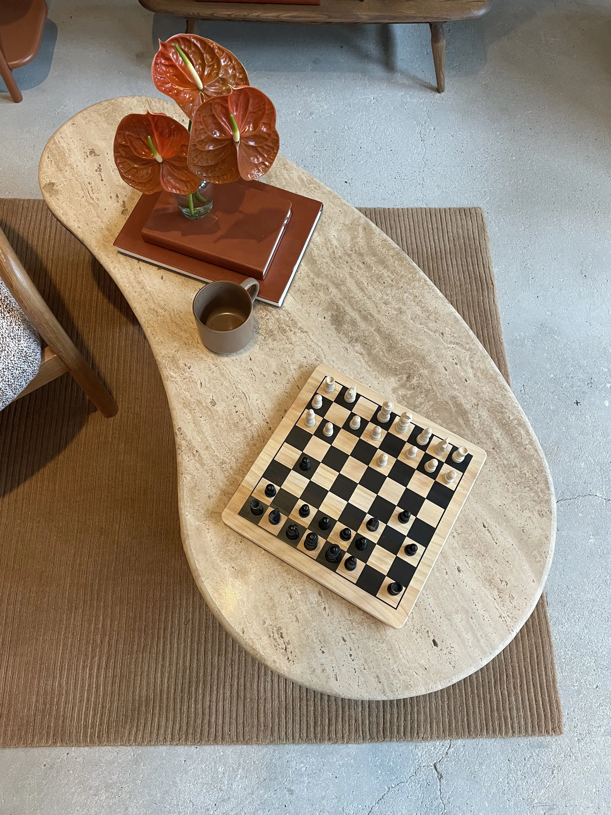 A wooden chessboard with chess pieces in starting positions, placed on a light-colored oval marble table. There is a gray mug, a closed notebook, a reddish-brown notebook, and a decorative plant with reddish-orange leaves in a glass vase on the table