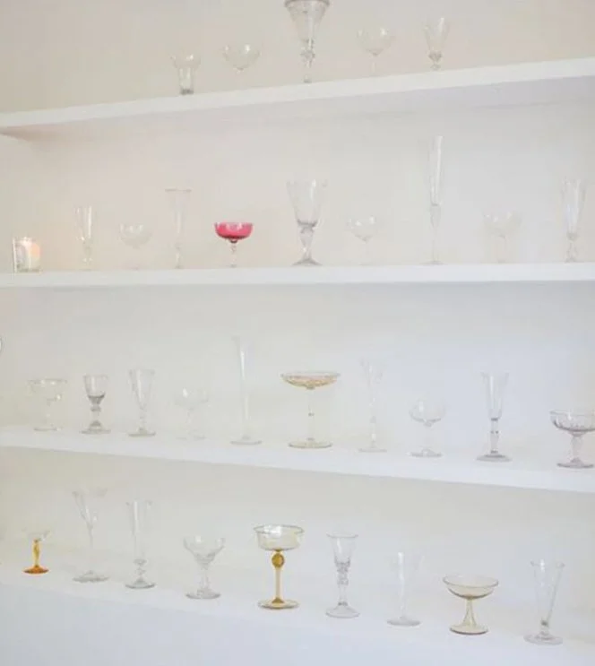 Display of variously shaped and colored glassware on white shelves against a white wall.