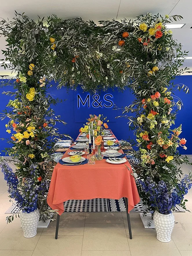 A decorated dining table with a coral tablecloth, set with plates, glasses, and yellow bowls, positioned under a floral arch with greenery, flowers, and small oranges. Blue Vases with purple flowers are at the base of the arch, and a blue wall with t