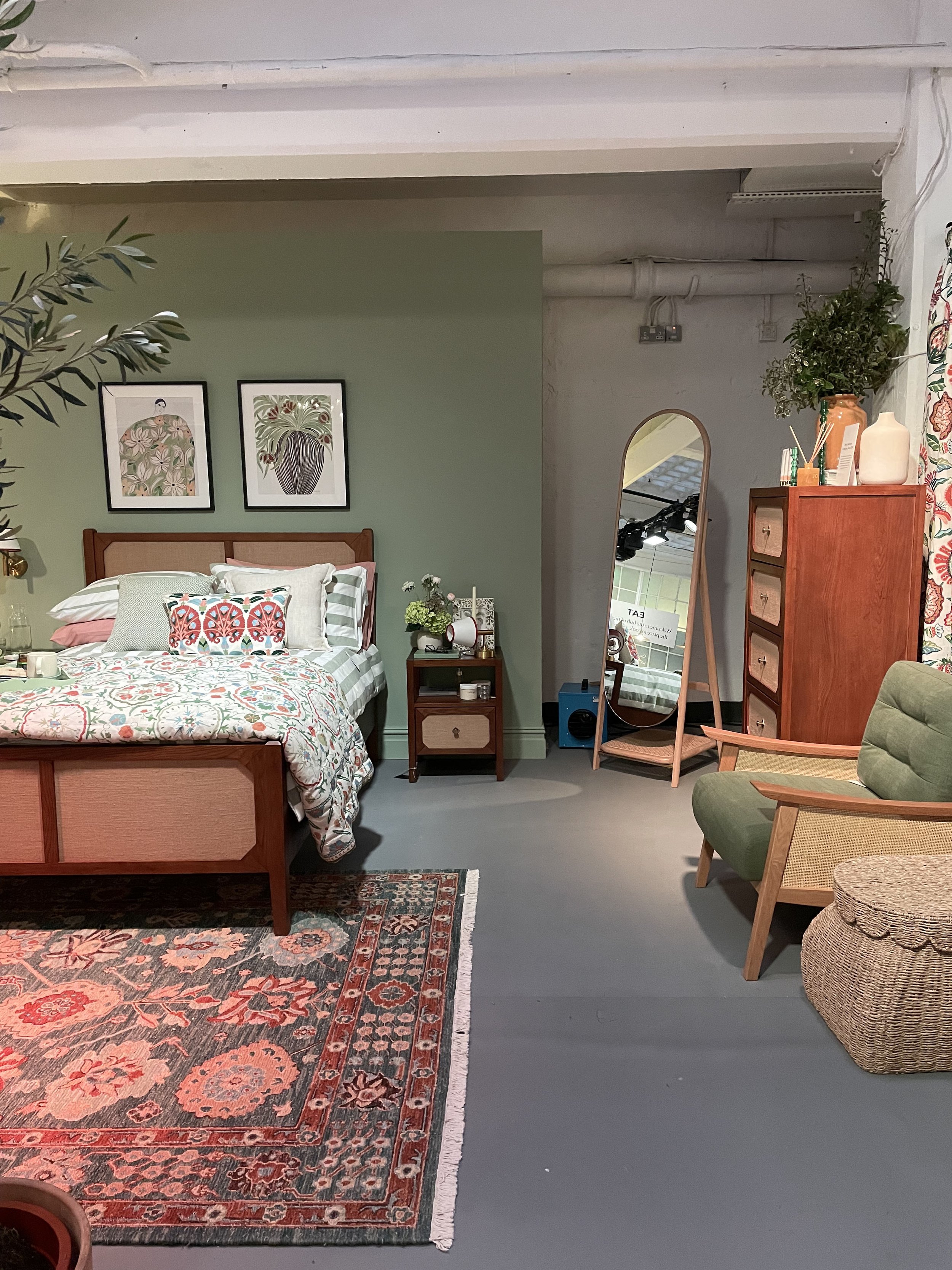 A cozy bedroom with a wooden bed and colorful bedding, a bedside table with flowers and books, a tall wooden dresser with decor, a large mirror with a wooden frame, a green armchair, a woven basket, a patterned area rug, and framed botanical art on t