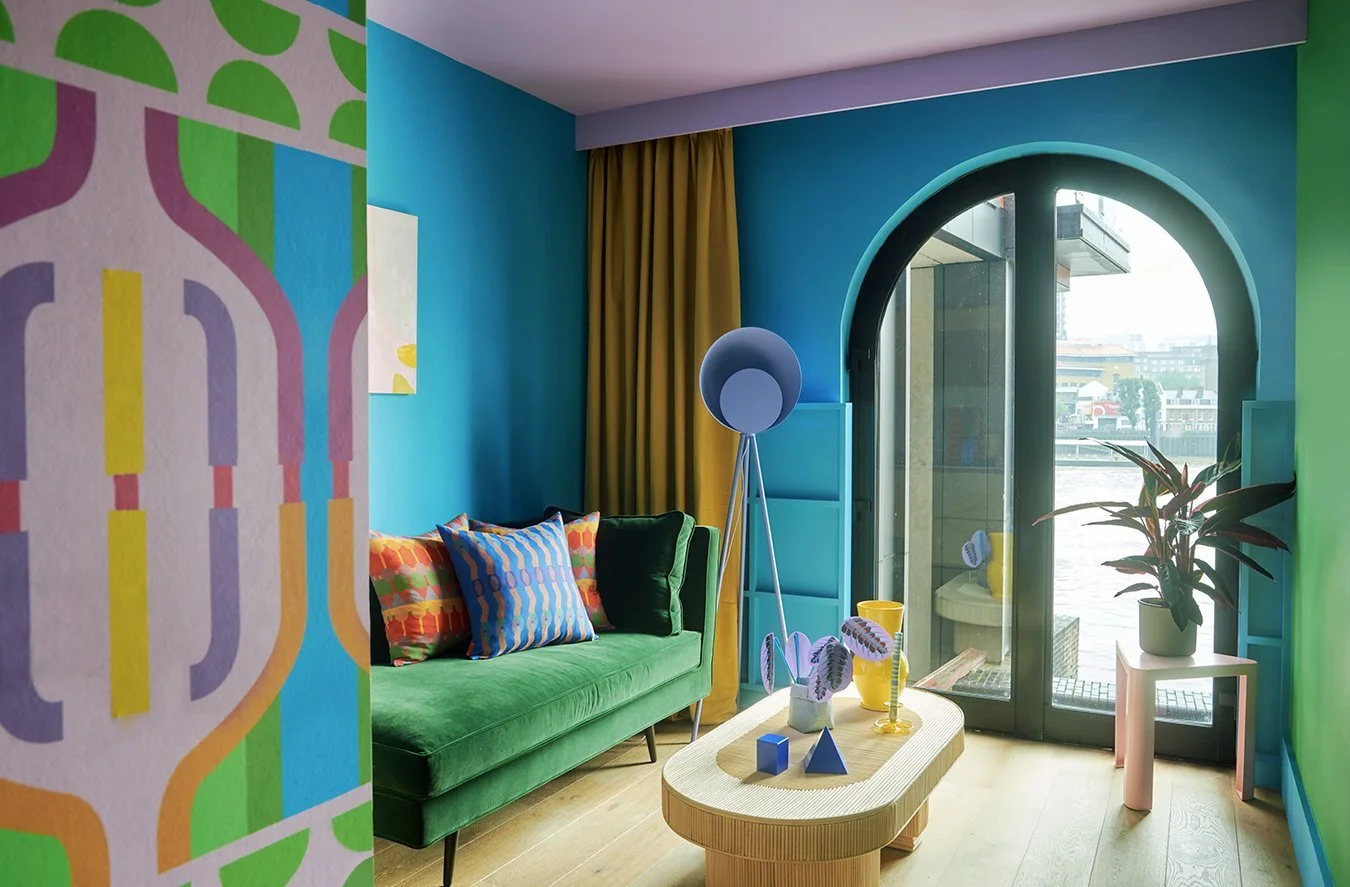 Bright living room with blue and green walls, colorful cushions, and a green sofa. A round wooden coffee table with geometric décor, a floor lamp, and a potted plant by a glass door leading outside. Gold curtains partially covering the door, with cit