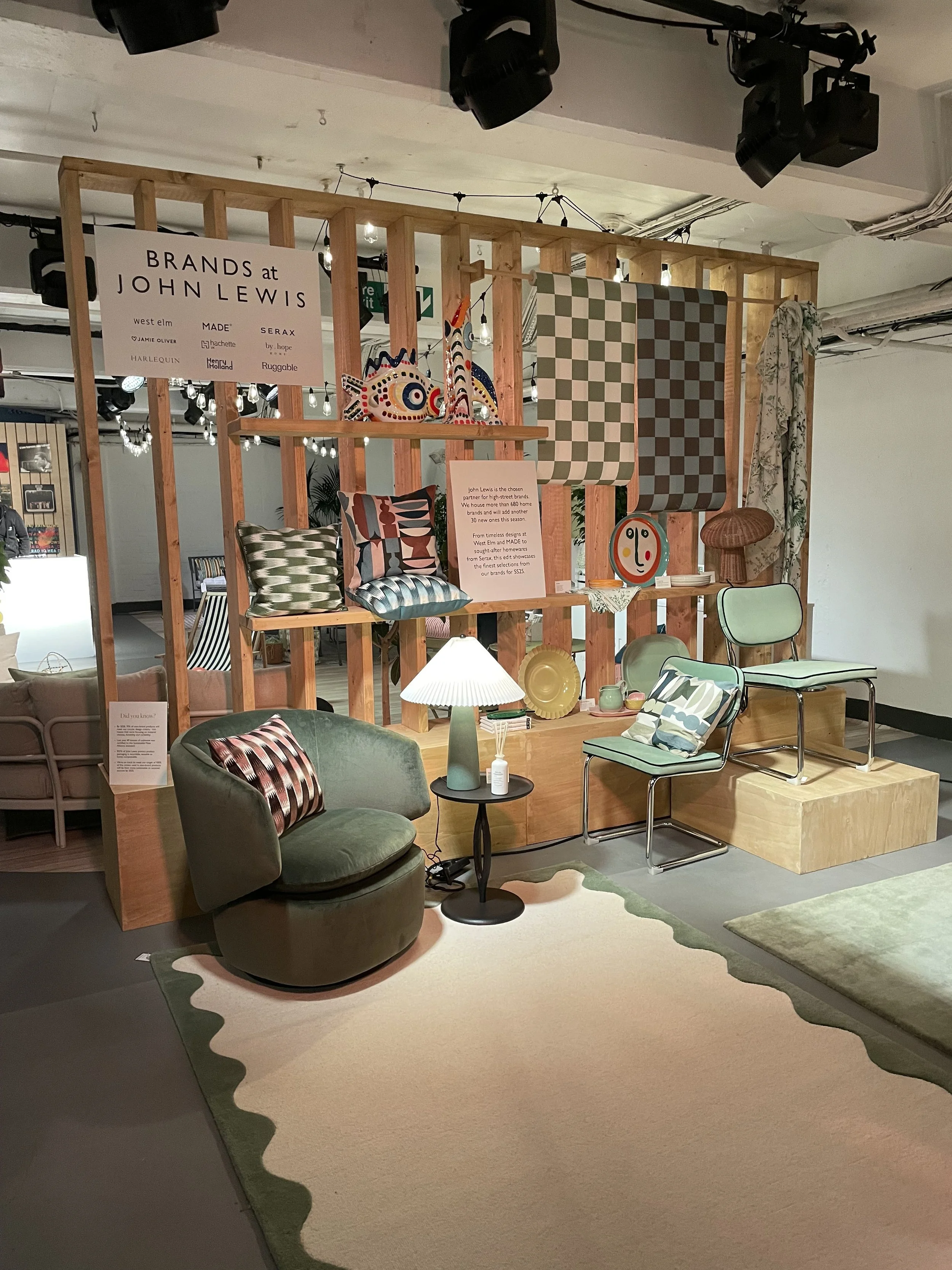 Display of home decor items and furniture, including chairs, pillows, lamps, and artwork, with a wooden pallet backdrop and a sign that says "Brands at John Lewis."