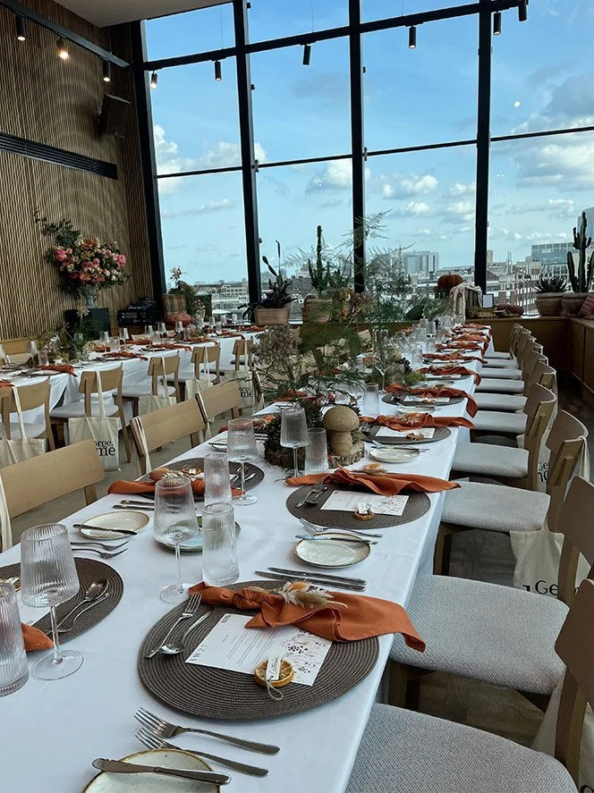 Long dining table set for a formal event with orange napkins, glassware, plates, and silverware, inside a modern room with large windows overlooking a cityscape and sky.