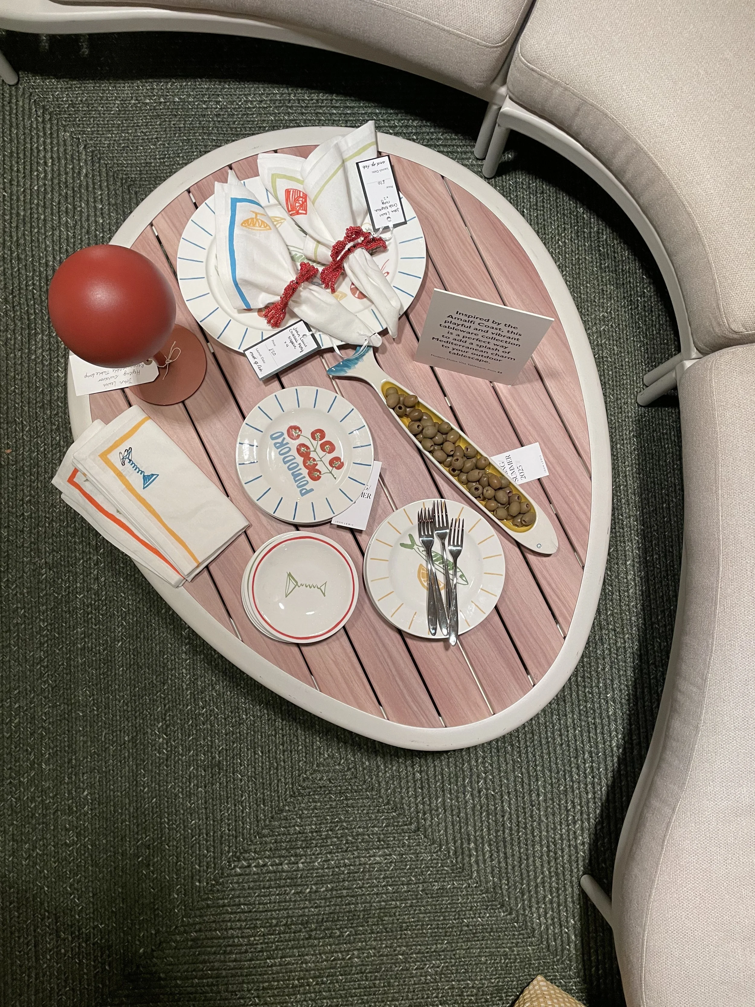 A table setting inspired by the movie 'A Average.' It features a plate with cherry tomato design, a small bowl, a platter of green olives, colored napkins, a red sphere decor, and silverware, all arranged on a pink wooden table with surrounding beige