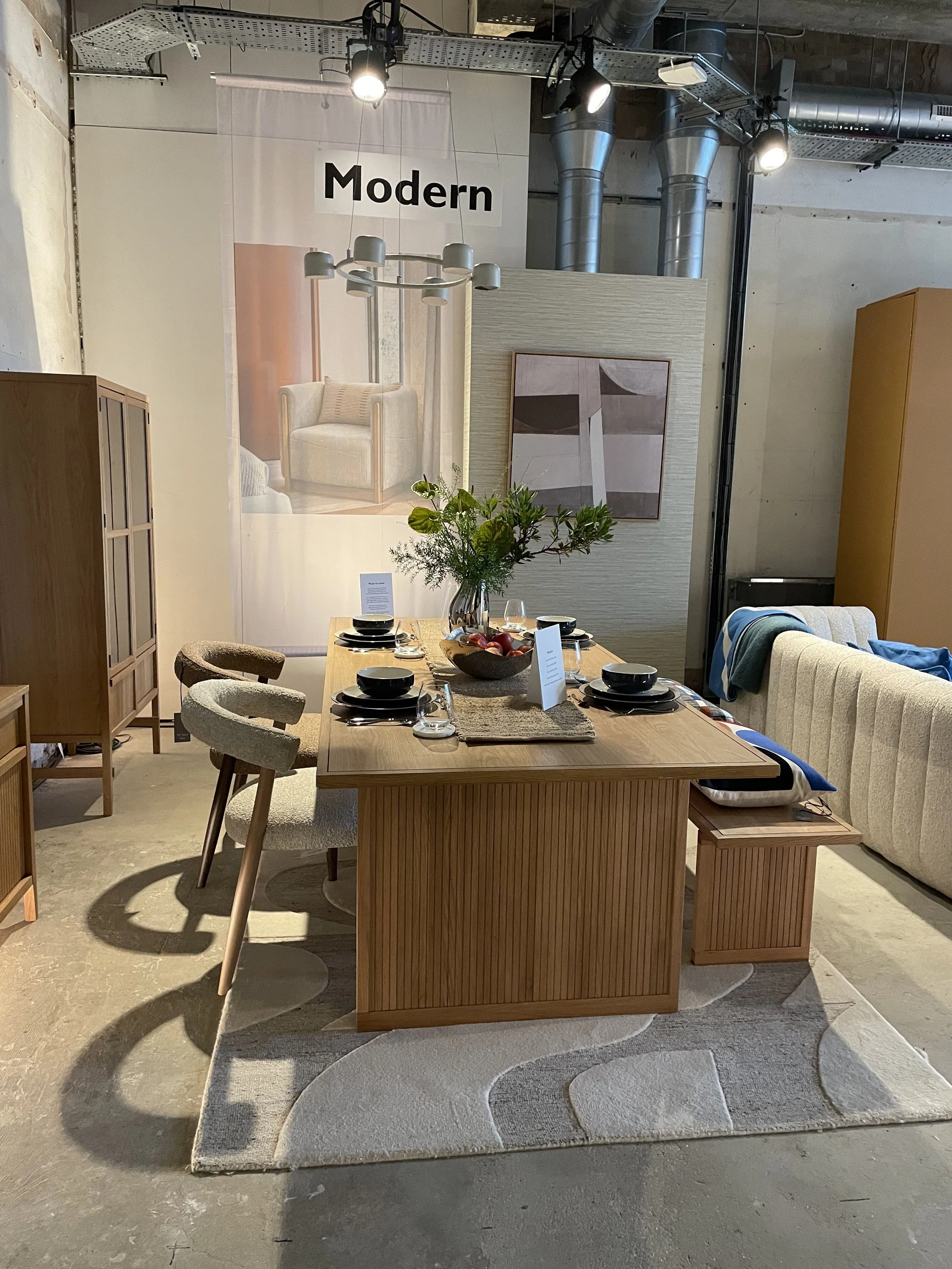 Modern dining room with a wooden table set for four, black dinnerware, a centerpiece with green foliage, surrounded by various chairs and a sofa, with a large banner reading 'Modern' and abstract art on the wall.