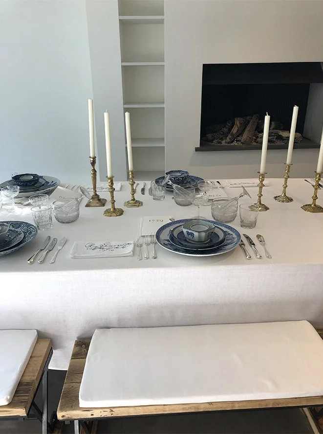 Dinner table set with blue and white china, gold candlesticks with white candles, glassware, silverware, a cloth napkin, and a small ceramic dish, in front of a fireplace and empty built-in shelves.