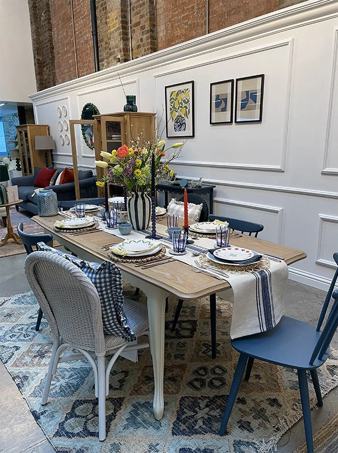 A dining table set with plates, glasses, napkins, and a centerpiece of colorful flowers, in a room with a white paneled wall, artwork, and wooden shelving with decor.