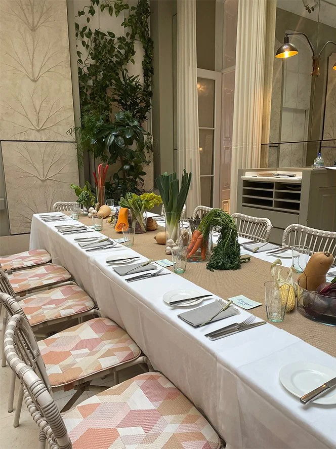 A long dining table set for a meal, with a white tablecloth, place settings, and a centerpiece with vegetables and plants, in an elegant room with tall windows and draped curtains.