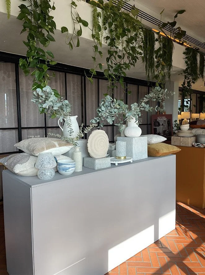 Decorative display with white vases, bowls, and cushions on a gray table, accented by hanging green plants inside a room with a black-framed glass wall.