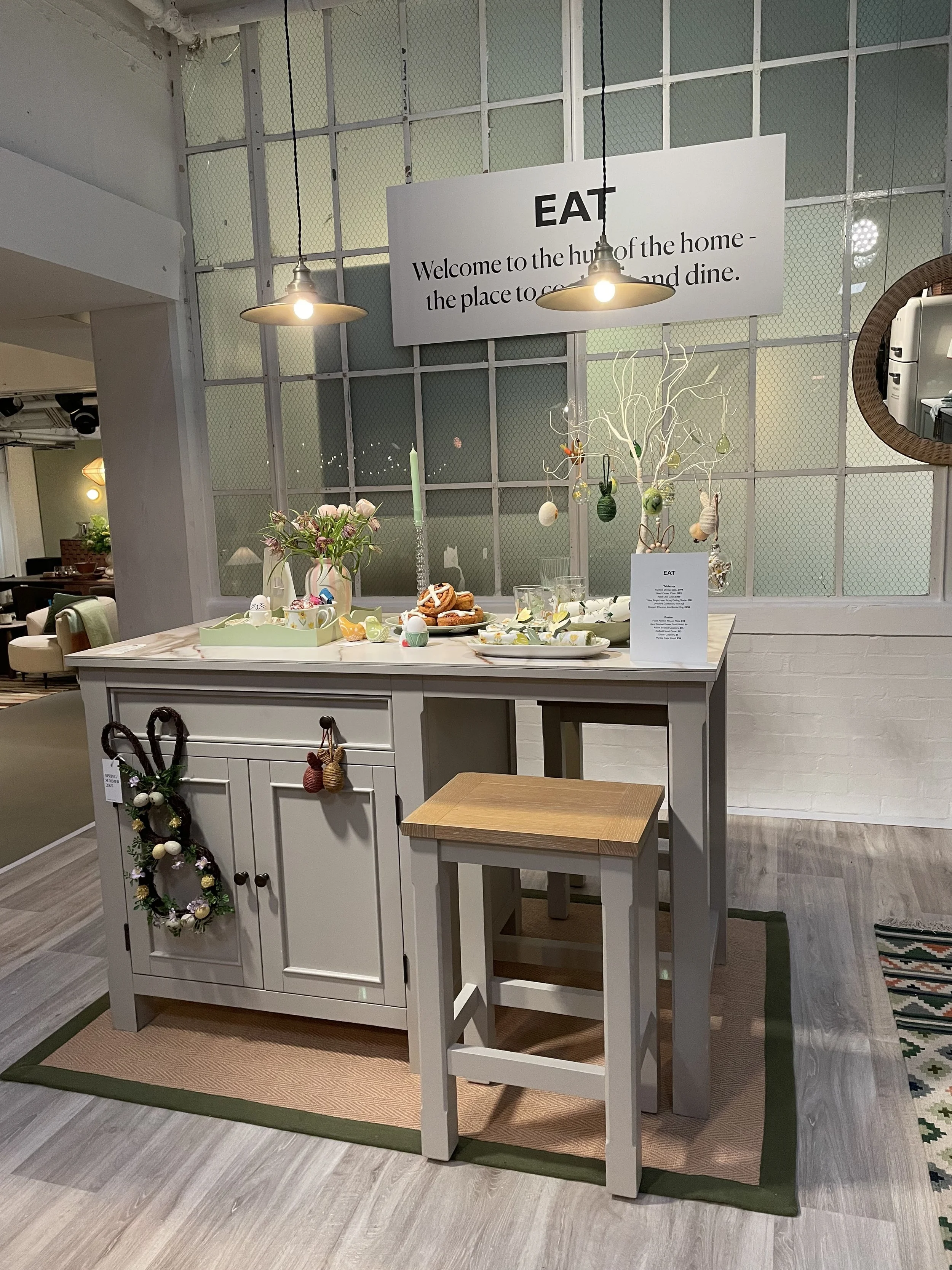 Decorative table display with Easter-themed decorations, flowers, and treats, including hanging eggs and a wreath, inside a home decor store.