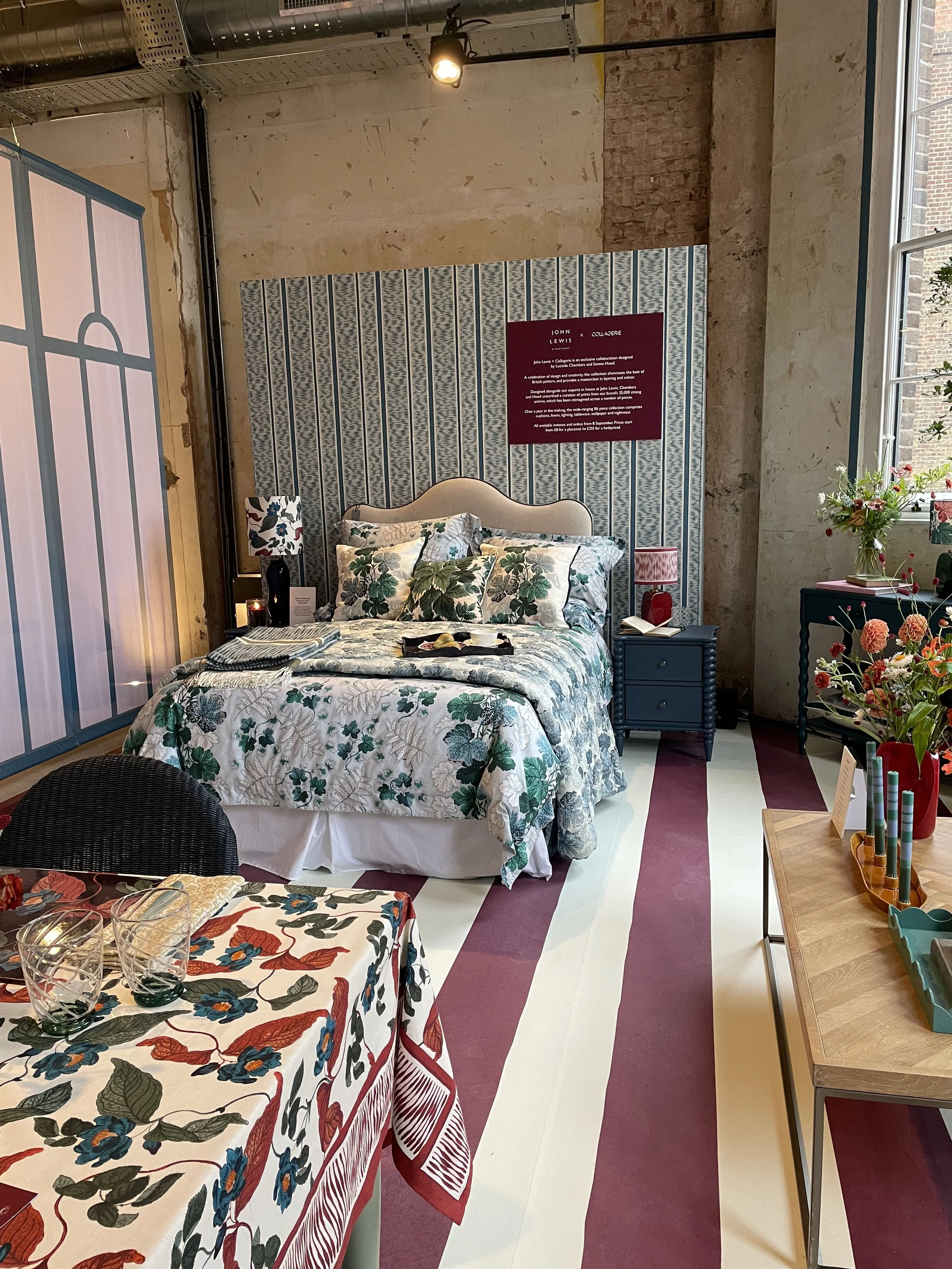 A bedroom display in a store, featuring a bed with floral bedding, decorative pillows, side tables with lamps, a window with natural light, and various decorative items and furniture.