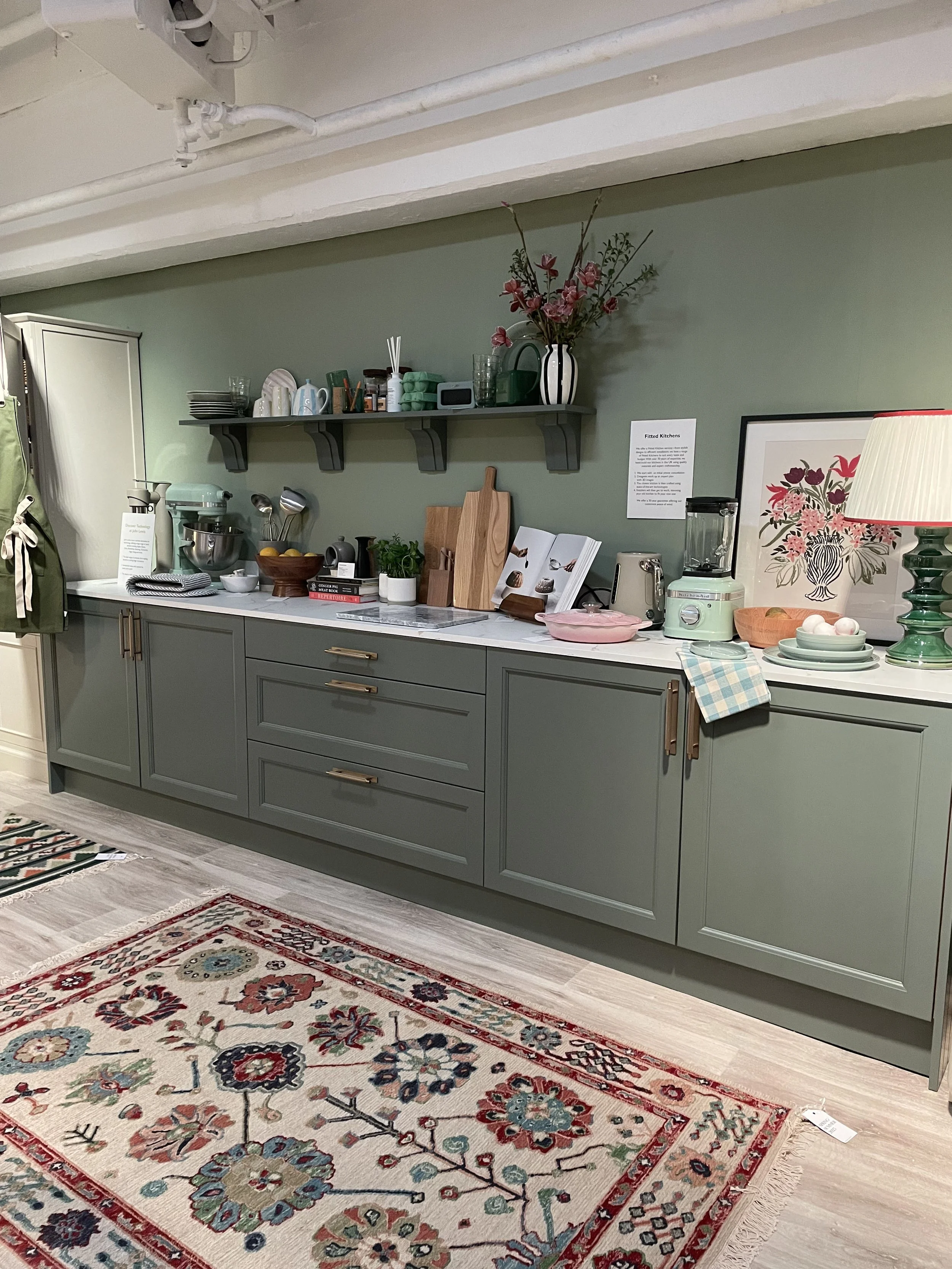A kitchen with green cabinets, a white countertop, and a decorative patterned rug on the floor. The countertop has various kitchen items, including cutting boards, a blender, books, and dishes. A gray shelf above holds dishes, a vase with pink flower