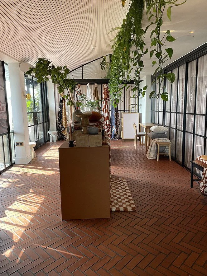 Indoor space with brick flooring, large windows, hanging plants, and various furniture including chairs, a small table, and clothing or fabric displayed in the background.