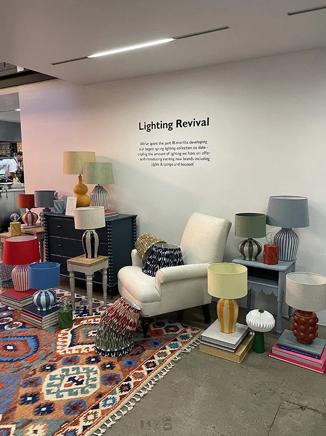 Interior display with colorful lamps, vases, and furniture pieces arranged on tables and a patterned rug, with a wall sign titled 'Lighting Revival'.