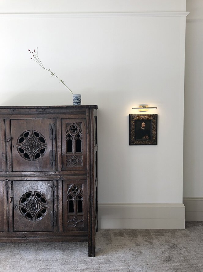 A dark wooden cabinet with intricate circular and floral carved designs, a small framed portrait on the white wall beside it, and a tall, thin vase with a single long, wispy branch on top of the cabinet. The room has light-colored carpet and a plain 