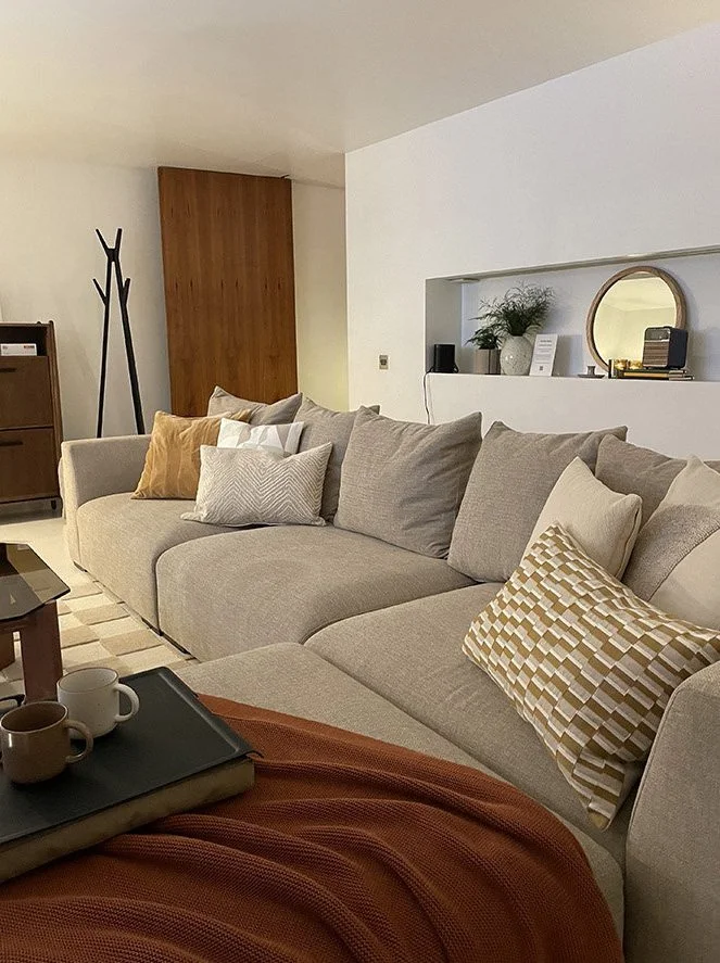 Living room with a large beige sectional sofa and various throw pillows, a coffee table with cups and a tray, a sideboard, and decorative items on a built-in shelf.
