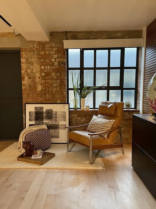 Cozy living room corner with a mustard armchair, a patterned pillow, a large framed abstract black and white artwork, a small wooden tray with a cup, books, and decorative objects, a potted plant on the window sill, and a brick wall backdrop with a l
