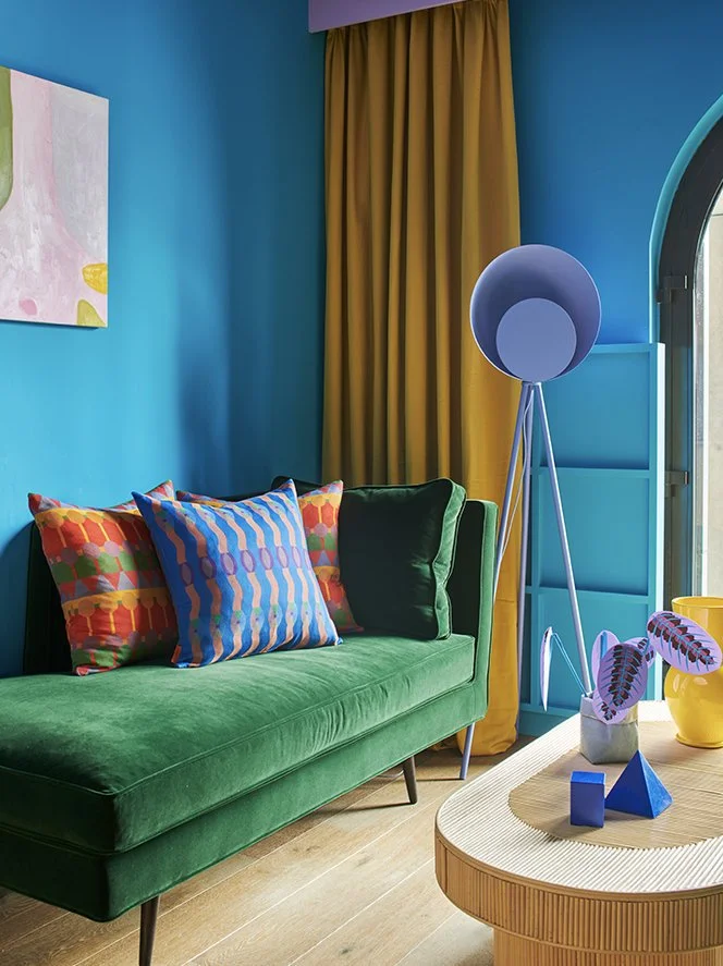 A living room with a vibrant green velvet sofa adorned with colorful, patterned pillows. A yellow curtain hangs in front of a window, with a blue wall behind. A unique blue floor lamp stands next to the sofa, and a wooden coffee table with decorative