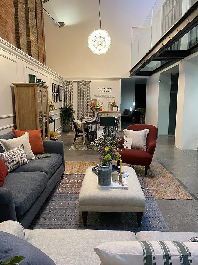 Interior of a living and dining room with sofas, armchairs, a coffee table with vases and books, a dining table with chairs, and decorative elements like curtains, plants, and artwork.