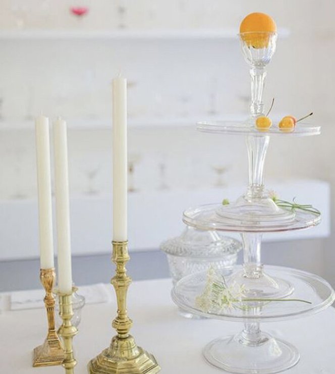 Decorative display with tall white candles in gold holders and a tiered glass stand holding cherries and an orange.