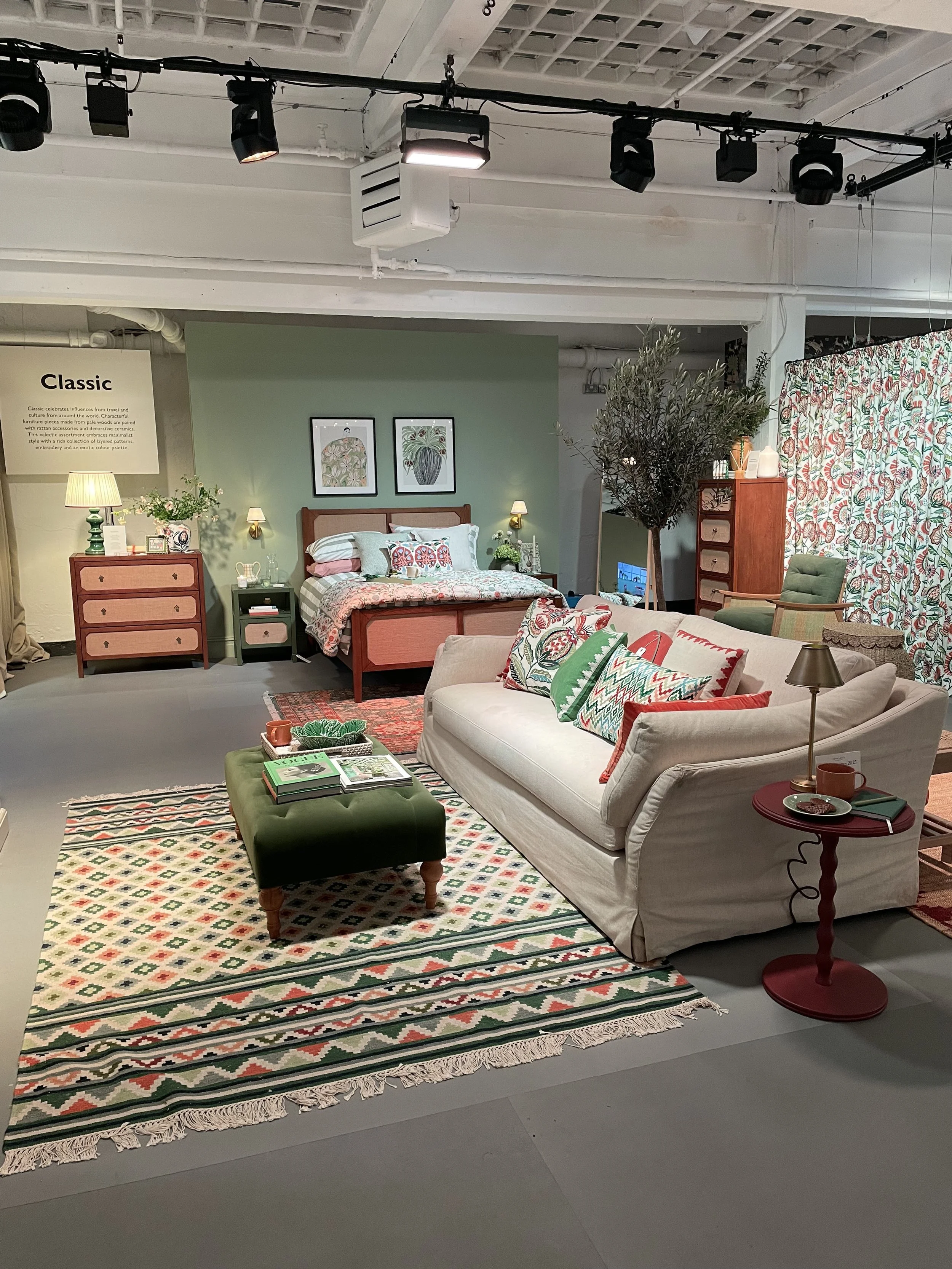 A bedroom and living room setup with a green accent wall, a bed with patterned pillows, a sofa with colorful pillows, a patterned rug, a green ottoman, and various furniture pieces and decor.