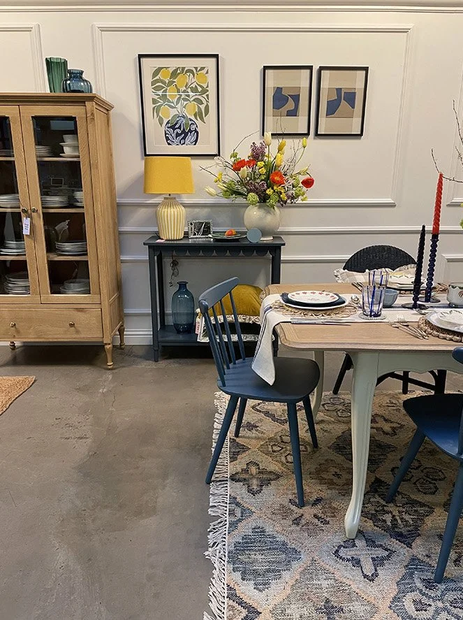 A dining room scene with a striped blue table, dark blue chairs, a floral centerpiece, and a yellow table lamp. There are three framed abstract art prints on the wall, and a light wooden china cabinet filled with dishes.