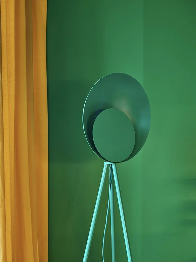 Modern floor lamp with a circular design against a green wall, next to yellow curtains.
