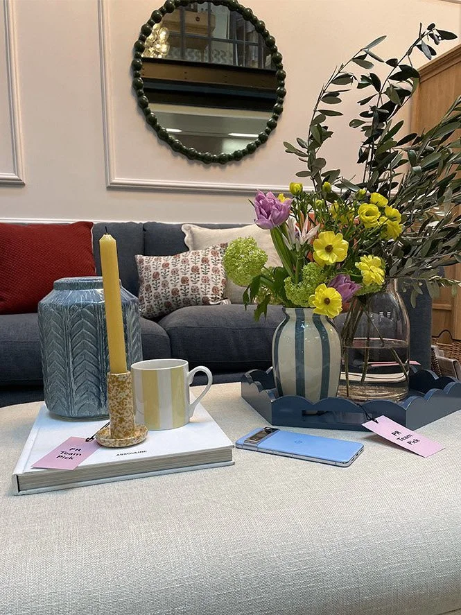A living room coffee table with a large striped vase of colorful flowers, a blue patterned vase, a yellow candle in a holder, a mug, a smartphone, and pink gift tags. In the background, there is a gray sofa with red and patterned pillows, a round mir