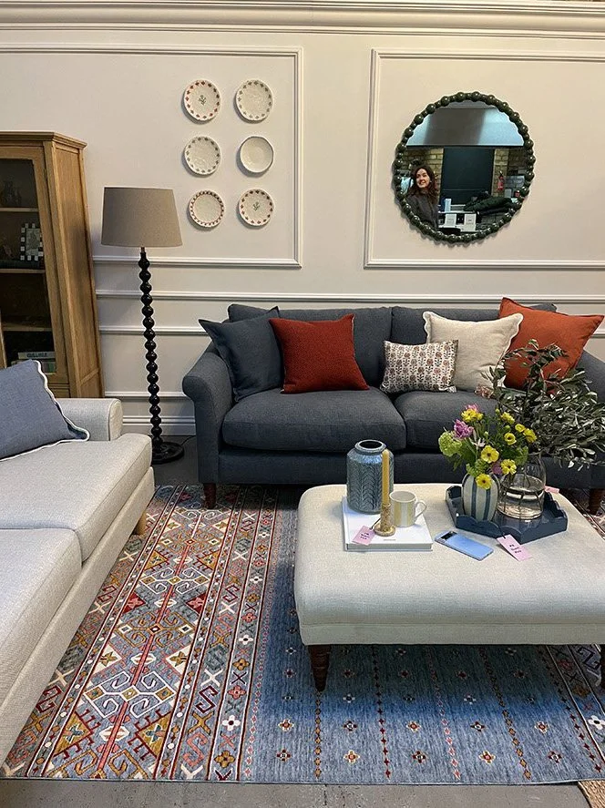 A cozy living room with a dark gray sofa, colorful throw pillows, a white tufted ottoman with a tray holding a vase of flowers, a mug, and a remote. A patterned area rug covers the floor. A tall floor lamp stands beside a light-colored armchair. Wall