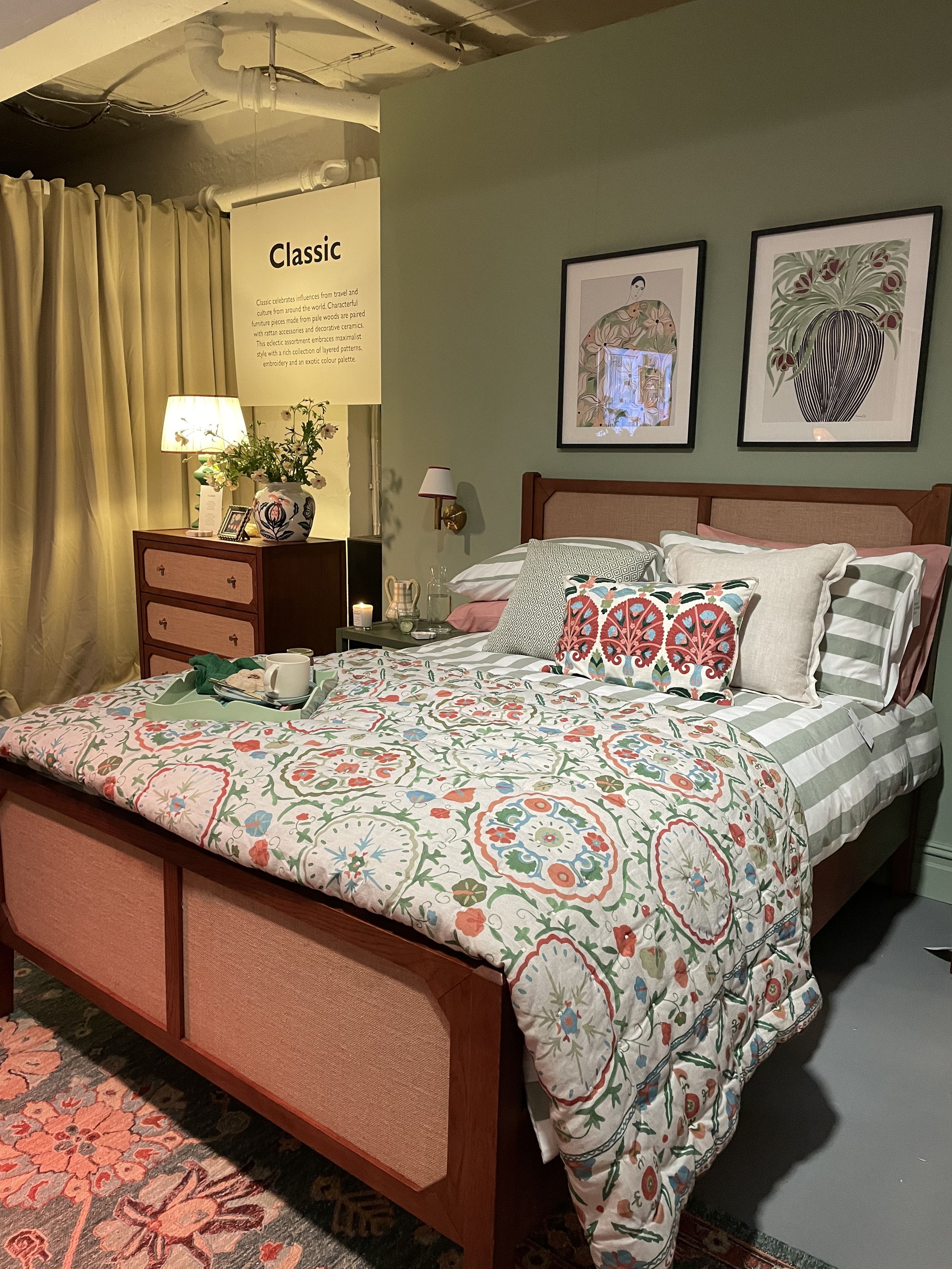 A neatly made bed with floral patterned bedding and multiple decorative pillows. There are framed pictures above the bed, a wooden dresser with a table lamp, a pot of flowers, and a sign labeled "Classic." The room has soft lighting and a cozy, decor