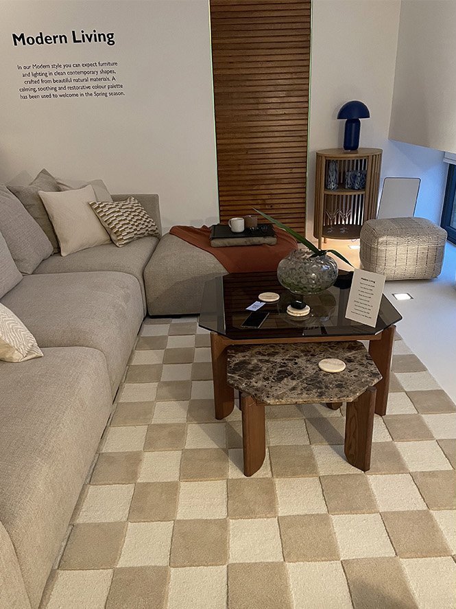 Living room with a beige sectional sofa, decorative pillows, and a checkered beige and cream rug. A black coffee table with a marble accent table underneath, holding a large glass vase with a plant, a menu, and other small objects. In the background,