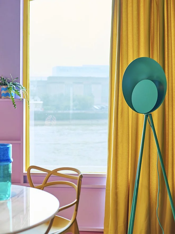 Bright interior space with pink wall, large window with yellow curtains, a green floor lamp, a portion of a white table with blue jars, and a wooden chair.